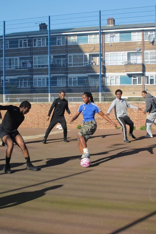 A five-a-side game in a fenced cage beneath a block of flats